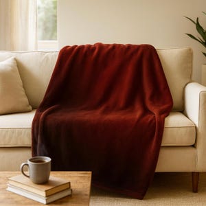 May include: A deep burgundy blanket with a soft texture, draped over a beige sofa. The blanket has a gradient effect, fading to a darker shade. A grey mug and books are on a wooden table in front of the sofa. The room has a warm, inviting atmosphere.