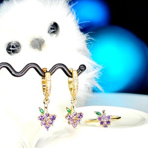May include: Gold-toned hoop earrings and a matching ring, each adorned with a cluster of purple and clear gemstones, resembling grapes. The earrings hang from a black display stand, with a white fluffy background and blurred blue and pink orbs.