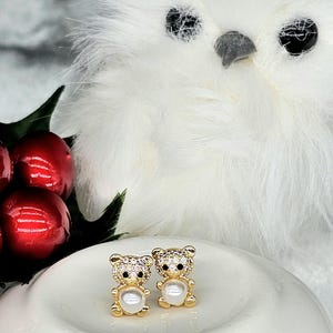 May include: A pair of gold-colored bear-shaped earrings with clear crystal accents and a pearl belly. The earrings are displayed on a white ceramic surface, with a white owl-like figure in the background.