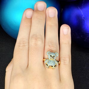May include: A gold-colored ring featuring a teddy bear design. The bear is adorned with light blue gemstones, and the ring has a simple band. The ring is shown on a hand, with a blurred background of blue and purple ornaments.