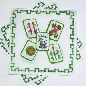 May include: A square white paper napkin featuring a green bamboo-style border. The center displays four Mahjong tiles arranged around a central Joker tile, which depicts a panda holding a bamboo stalk. The other tiles show bamboo and floral motifs.