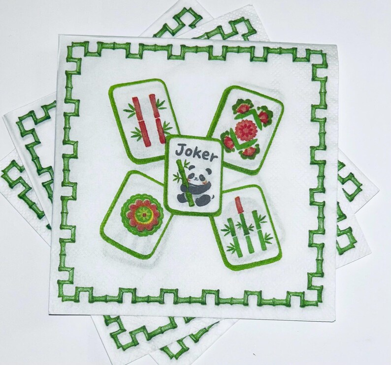 May include: A square white paper napkin featuring a green bamboo-style border. The center displays four Mahjong tiles arranged around a central Joker tile, which depicts a panda holding a bamboo stalk. The other tiles show bamboo and floral motifs.
