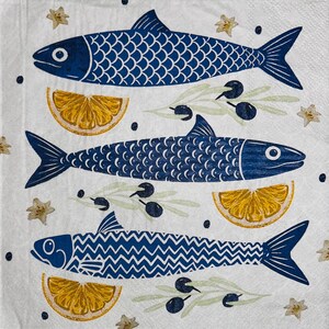 May include: White paper napkin with a blue fish design. The design includes three blue fish with detailed scales, olive branches, lemon slices, and star-shaped accents. The fish are arranged vertically on the napkin.