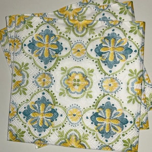 May include: White paper napkins with a repeating pattern of teal, yellow, and green floral designs. The napkins are folded and stacked, showcasing the detailed print. The design features stylized flowers and geometric shapes.
