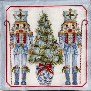 May include: A decorative square napkin featuring a Christmas theme. Two blue and white nutcrackers flank a decorated Christmas tree in a blue and white vase. The napkin has a red border and a light blue background.