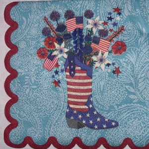May include: A decorative, patriotic-themed kitchen towel. The design features a cowboy boot with the American flag, filled with flowers and small American flags. The background is a light blue paisley pattern with a red scalloped border.