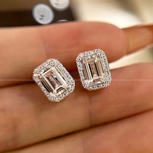 May include: A pair of square emerald-cut stud earrings. Each earring features a large, clear, rectangular gemstone surrounded by a halo of smaller, sparkling stones. The earrings are set in a silver or white metal.
