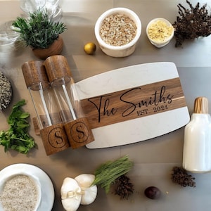 May include: A white and brown rectangular serving board with the text "The Smiths EST. 2024" engraved on the wood. The board has a white marble top and a brown wood bottom. Two glass salt and pepper shakers are attached to the board.