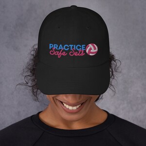 May include: Black baseball cap with the text "PRACTICE Safe Sets" in pink and blue with a pink and white volleyball graphic.