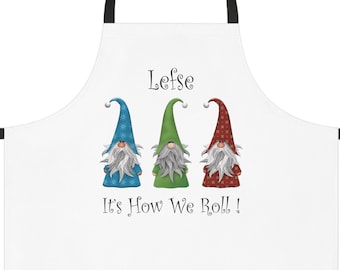 Puede incluir: Delantal blanco con una correa ajustable negra. El delantal tiene un diseño de tres gnomos con sombreros azules, verdes y rojos. El texto "Lefse It's How We Roll!" está impreso en el delantal.