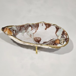 May include: A decorative oyster shell dish featuring a marbled white and brown interior with a gold-painted rim. The shell is supported by a small gold metal stand and is adorned with two small, textured seashells attached to the inner edge.