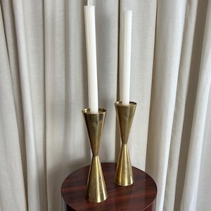 May include: Two brass candle holders with white candles are displayed on a dark wood pedestal. The candle holders have a unique hourglass shape. The background features a neutral-colored curtain.