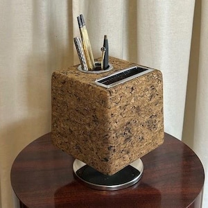 May include: A cube-shaped desk organizer made of cork, with a silver-colored trim and base. The organizer holds pens and pencils. The desk accessory sits on a dark wood table, against a neutral-colored curtain backdrop.