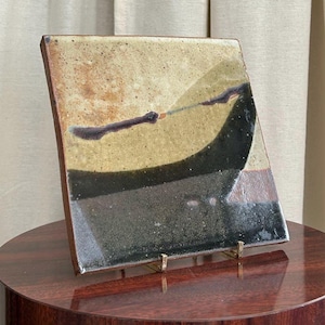 May include: A square ceramic art piece with a unique abstract design. The artwork features a blend of colors, including beige, black, brown, and gold. The piece is displayed on a dark wooden pedestal, showcasing its artistic qualities.