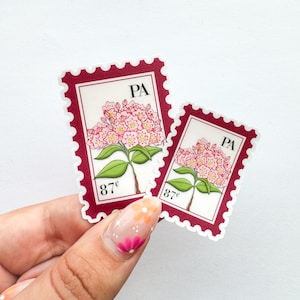 Puede incluir: Dos pegatinas con forma de sellos postales. Los sellos tienen un fondo blanco con una ilustración de flores rosas, hojas verdes y el texto "PA" y "87¢". Los sellos tienen un borde rojo oscuro.