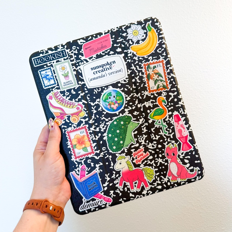 Op de afbeelding: Een zwart-witte compositienoteboek met verschillende kleurrijke stickers, waaronder een rolstoel, een flamingo, een egel, een boekenworm, een bloem, een lavalamp, een eenhoorn en een dinosaurus. Het notitieboek heeft de woorden "BOOKISH", "Mistakes", "sunspoken creative (amanda's version)", "book worm" en "demure" erop geschreven.