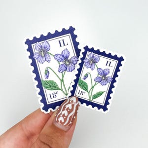 Può includere: Due adesivi che assomigliano a francobolli vintage. Ogni francobollo presenta un'illustrazione dettagliata di viole viola con foglie e steli verdi. I francobolli hanno un bordo blu scuro e il testo "IL" e "18¢".