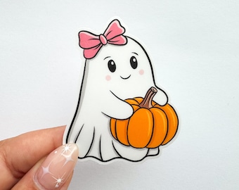 Puede incluir: Adhesivo de fantasma blanco con un lazo rosa sosteniendo una calabaza naranja. El fantasma tiene una cara sonriente con mejillas rosadas. Diseño de estilo de dibujos animados lindo, perfecto para Halloween.