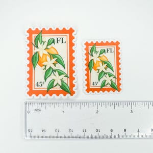 Op de afbeelding: Twee postzegels met sinaasappelbloesem met de staat afkorting "FL" en de prijs "45&cent;" erop gedrukt. De postzegels zijn op een witte achtergrond met een liniaal voor schaal.