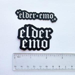 Puede incluir: Dos pegatinas en blanco y negro con el texto "elder emo" en fuente g&oacute;tica. Una es rectangular, la otra es un rect&aacute;ngulo redondeado. Una regla est&aacute; colocada debajo, midiendo en cent&iacute;metros.