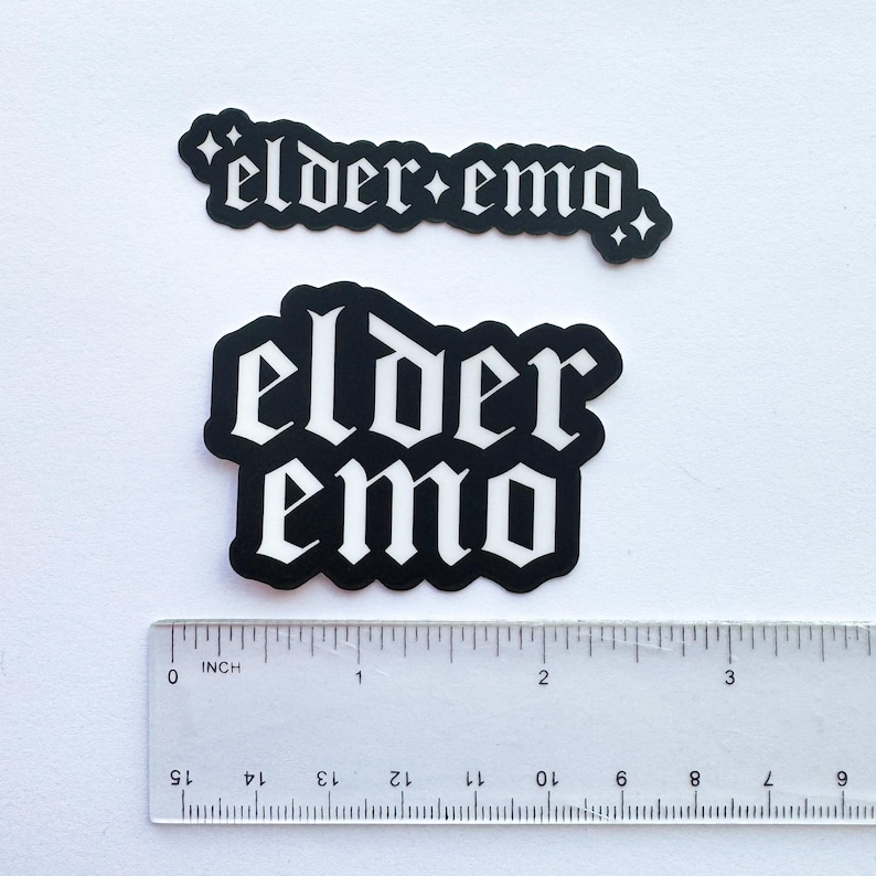 Puede incluir: Dos pegatinas en blanco y negro con el texto "elder emo" en fuente g&oacute;tica. Una es rectangular, la otra es un rect&aacute;ngulo redondeado. Una regla est&aacute; colocada debajo, midiendo en cent&iacute;metros.