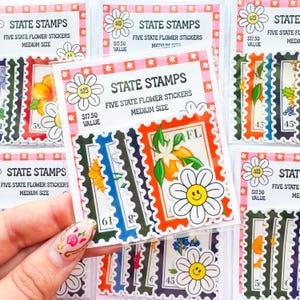 Op de afbeelding: Vijf vellen met stickers met ontwerpen van bloemen uit de staten. Elk vel heeft een andere bloem en staat-afkorting. De stickers zitten in een doorzichtige plastic zak met een roze en witte geruite achtergrond. De tekst op de zak luidt "State Stamps Five State Flower Stickers Medium Size".