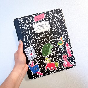 May include: A black composition notebook with various colourful stickers. The stickers include a bookworm, a hedgehog, a flamingo, a lava lamp, a unicorn, and a dragon. The notebook has the text "Composition Book" and "Mistakes" written on it.