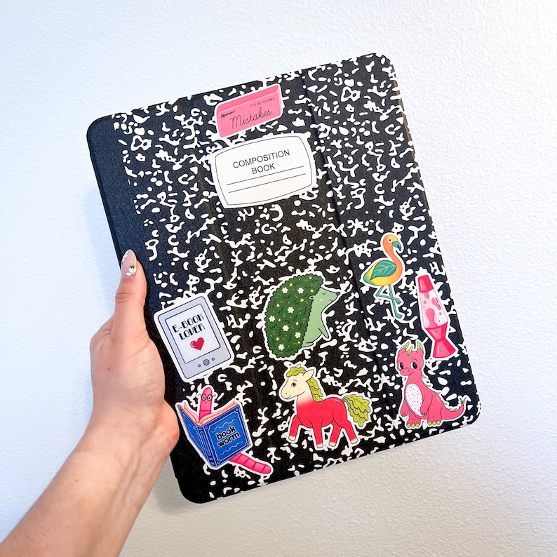 May include: A black composition notebook with various colourful stickers. The stickers include a bookworm, a hedgehog, a flamingo, a lava lamp, a unicorn, and a dragon. The notebook has the text "Composition Book" and "Mistakes" written on it.