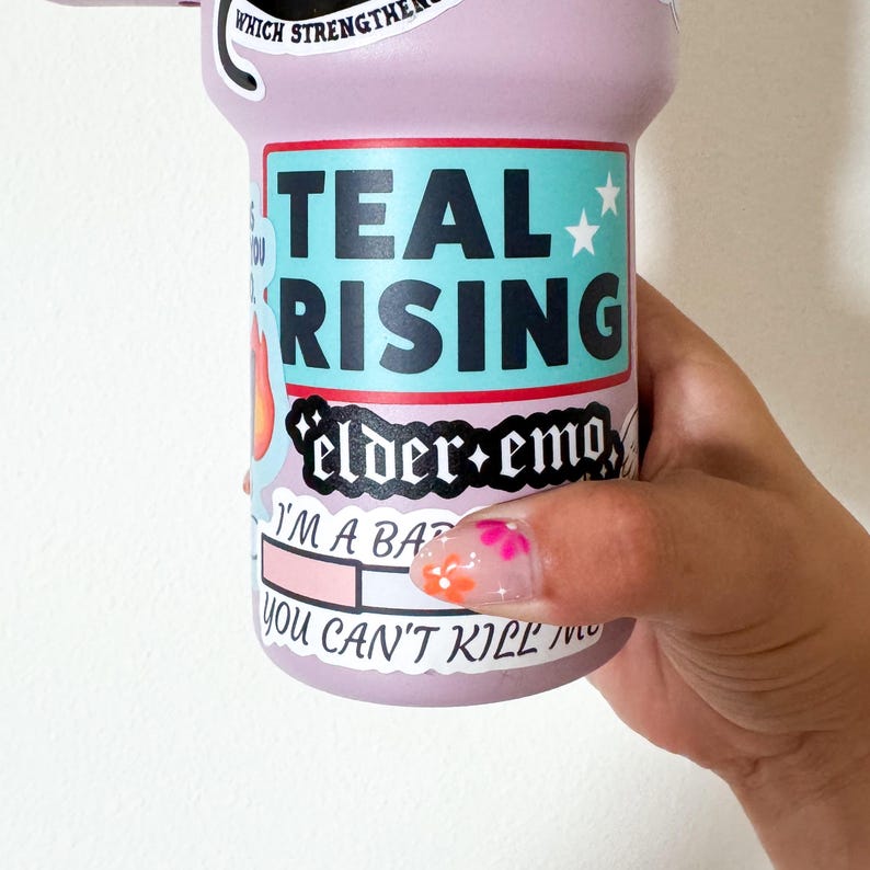 Puede incluir: Una botella de agua morada con las palabras "TEAL RISING" en turquesa y blanco. La botella tambi&eacute;n tiene las palabras "elder+emo" y "I'M A BA" impresas. La botella es sostenida por una mano con un dise&ntilde;o floral en las u&ntilde;as.