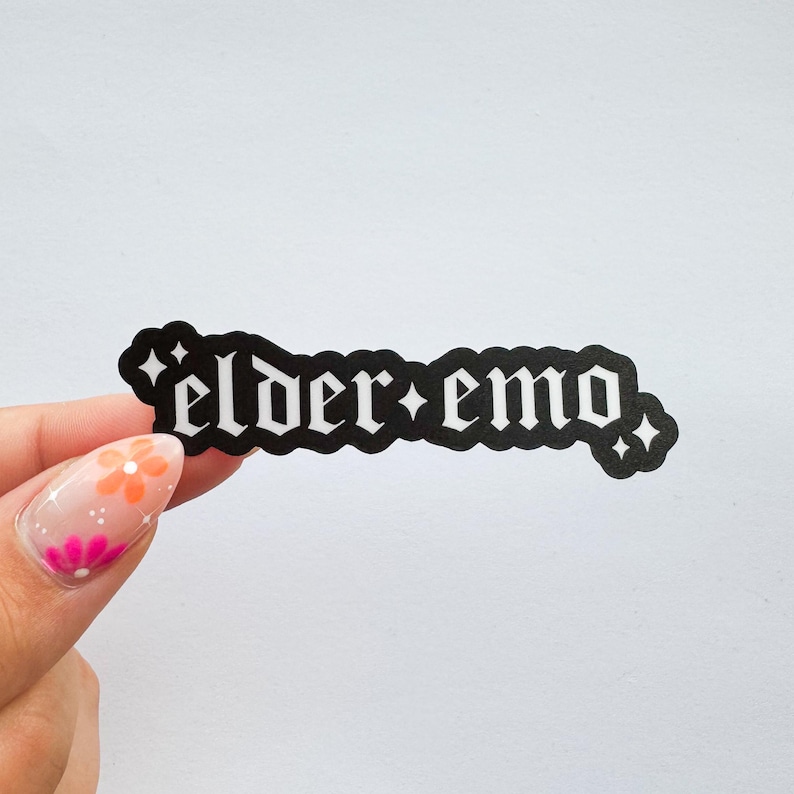 Puede incluir: Una pegatina negra con las palabras "elder emo" en una fuente g&oacute;tica. La pegatina tiene un borde blanco y detalles en forma de estrella. La pegatina est&aacute; sujeta por una mano con una manicura colorida.