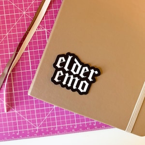 Puede incluir: Una pegatina en blanco y negro con la frase "elder emo" en fuente g&oacute;tica est&aacute; colocada sobre un cuaderno beige. Una esterilla de corte rosa con l&iacute;neas de cuadr&iacute;cula y una cinta est&aacute;n en el fondo.