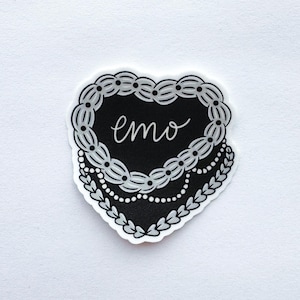 May include: A heart-shaped sticker with a black center and the word "emo" in white script. The heart is outlined with a decorative white border, featuring a beaded design and leaf accents. The sticker is on a white background.