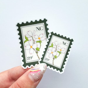 May include: Two stickers in a stamp design featuring white dogwood flowers with green leaves. The stickers have a dark green border and the text "NC" and "89&cent;". The stickers are held by a hand with a glittery nail design.
