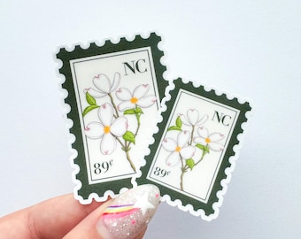 Op de afbeelding: Twee stickers in een postzegelontwerp met witte bloemen met groene bladeren. De stickers hebben een donkergroene rand en de tekst "NC" en "89¢". De stickers worden vastgehouden door een hand met een glitterende nageldesign.