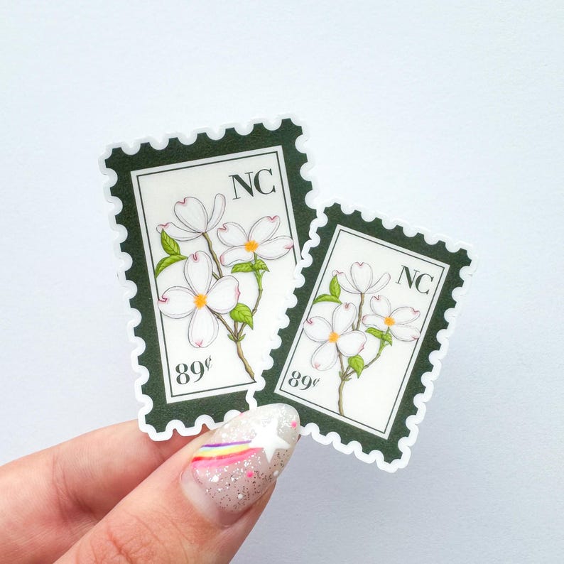 May include: Two stickers in a stamp design featuring white dogwood flowers with green leaves. The stickers have a dark green border and the text "NC" and "89&cent;". The stickers are held by a hand with a glittery nail design.