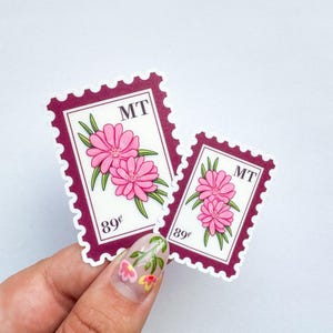 Puede incluir: Dos pegatinas con diseño de sello con flores rosas y hojas verdes. Los sellos tienen un borde burdeos y las letras "MT" en la parte superior. El precio "89¢" está impreso en la parte inferior.