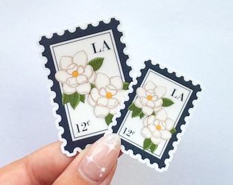 May include: Two stickers in the shape of postage stamps, featuring white magnolia flowers with yellow centers and green leaves. The stamps have a dark blue border and the text "LA" and "12¢".