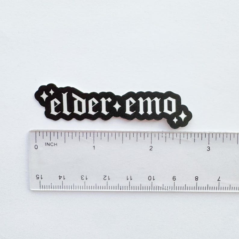 Puede incluir: Una pegatina negra con la frase "elder emo" en fuente g&oacute;tica, con detalles de estrellas. La pegatina est&aacute; junto a una regla, mostrando su tama&ntilde;o aproximado. La pegatina est&aacute; sobre un fondo blanco, midiendo unos 7.6 cm.