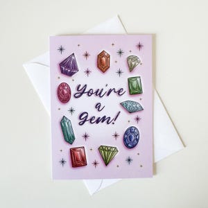 May include: A greeting card with a light pink background, featuring colorful gemstone illustrations and the text "You're a Gem!". The card is surrounded by small star illustrations and comes with a white envelope.