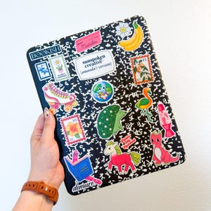 Puede incluir: Una cubierta de cuaderno de composici&oacute;n en blanco y negro decorada con pegatinas coloridas. Las pegatinas incluyen un pat&iacute;n, un pl&aacute;tano, un flamenco, una l&aacute;mpara de lava y el texto "BOOKISH". El cuaderno lo sostiene una persona que lleva un reloj marr&oacute;n.