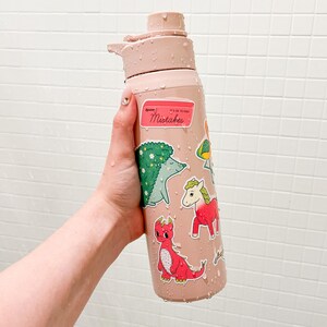 Puede incluir: Una botella de agua rosa claro adornada con pegatinas, incluyendo un erizo verde, un drag&oacute;n rojo y un unicornio rosa. La botella tiene una tapa abatible y el texto "Mistakes" en una pegatina.