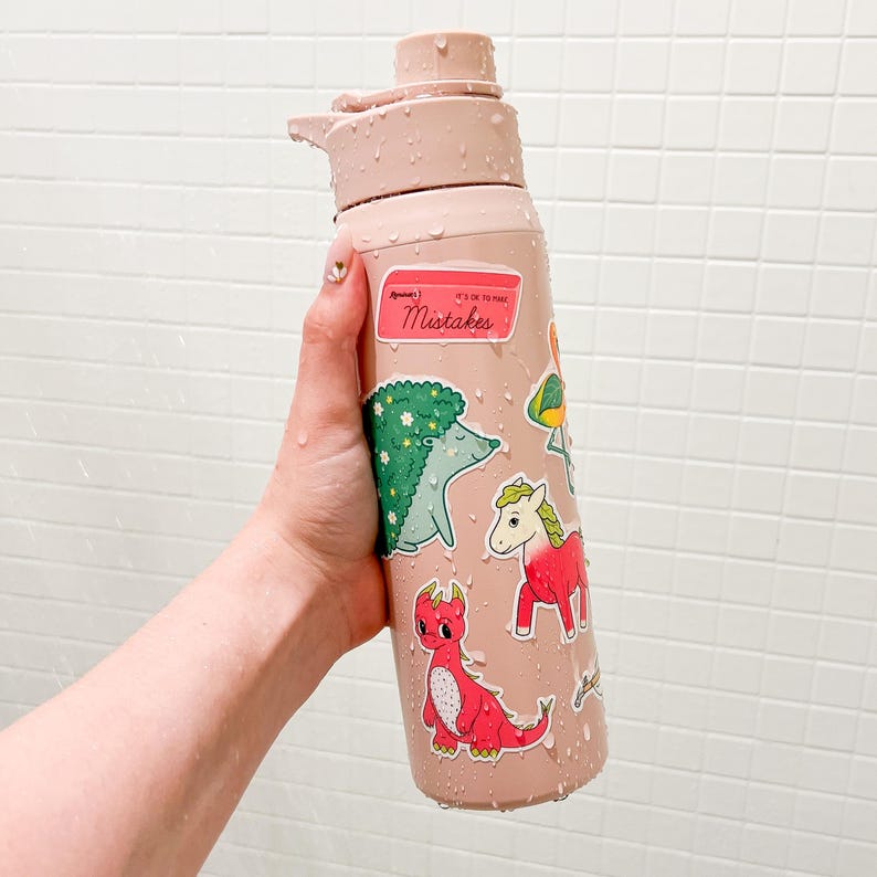Puede incluir: Una botella de agua rosa claro adornada con pegatinas, incluyendo un erizo verde, un drag&oacute;n rojo y un unicornio rosa. La botella tiene una tapa abatible y el texto "Mistakes" en una pegatina.
