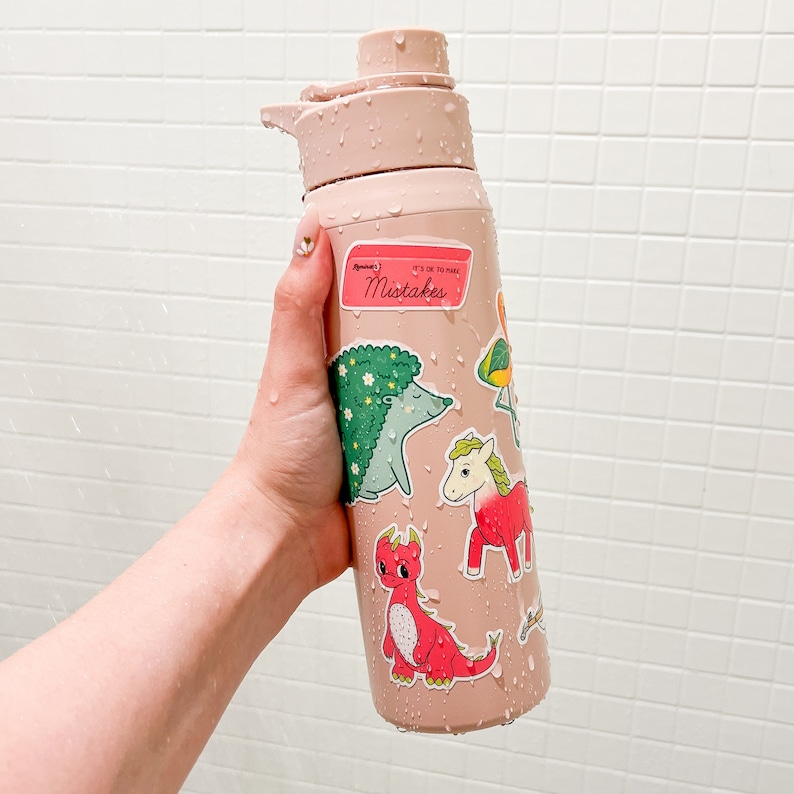 Op de afbeelding: Een roze herbruikbare waterfles met verschillende kleurrijke stickers met dieren en de tekst "It's OK to Make Mistakes".