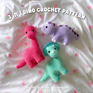 May include: Three crocheted dinosaur toys in pink, purple, and teal, with black eyes and white accents, are displayed on a floral patterned white surface. The text "3 in 1 DINO CROCHET PATTERN" is at the top. The bottom of the image reads "Digital PDF download: by crochetcalls."