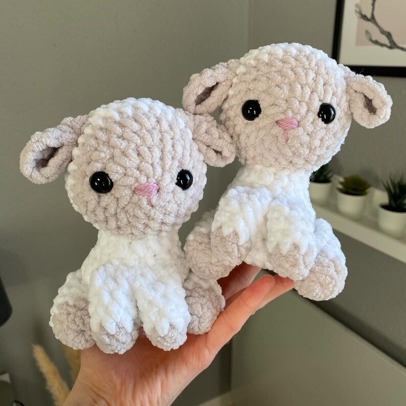 May include: Two crocheted plush lambs with white and beige fur. The lambs have black eyes, pink noses, and small, floppy ears.