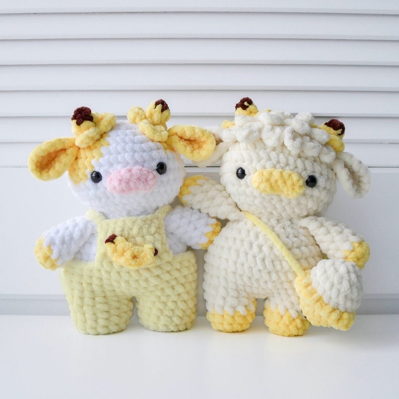May include: Two crocheted stuffed animals, one white with yellow accents and one yellow with white accents, are standing side-by-side. Both animals have a cow-like appearance with large eyes and a pink nose.