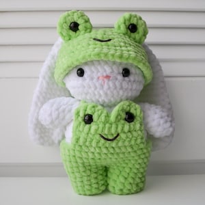 May include: A white crocheted bunny wearing a green crocheted frog hat and overalls. The frog hat has black eyes and a green smile. The overalls have black eyes and a green smile.