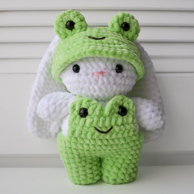 May include: A white crocheted bunny wearing a green crocheted frog hat and overalls. The frog hat has black eyes and a green smile. The overalls have black eyes and a green smile.