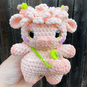 May include: A crocheted pink cow plush toy with a green strap and a light green leaf on its head. The toy has a pink nose and black eyes with gold glitter.