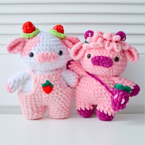 May include: Two crocheted stuffed cows, one pink and one white with pink accents, wearing overalls and holding strawberries.